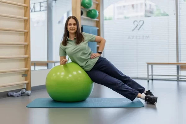 Kineziterapeutė atlieka šoninio stabilumo pratimą atsirėmusi į gimnastikos kamuolį