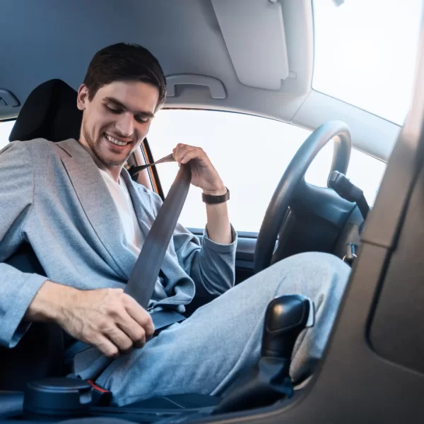 Automobilyje sėdintis asmuo segasi saugos diržą; matomas vairas, sėdynė, pavarų perjungimo svirtis ir automobilio salonas.