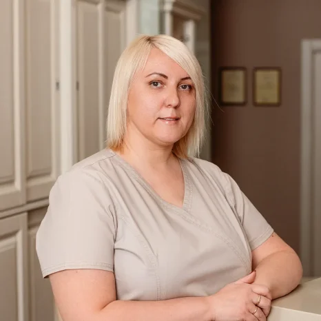 Odontologijos klinikos darbuotoja vilki šviesiai pilką medicininę aprangą ir stovi klinikos patalpose. Moteris žvelgia į kamerą, fone matomos šviesios spintelės ir koridorius