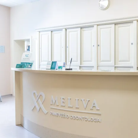 Šviesus odontologijos klinikos registratūros interjeras su baltomis spintelėmis ir registracijos stalu. Ant stalo matomas „Meliva Panevėžio odontologija“ logotipas