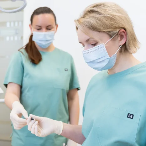 Dvi odontologijos klinikos specialistės su medicininėmis kaukėmis ruošiasi procedūrai. Viena jų rankoje laiko odontologinį instrumentą, o fone matoma šviesi klinikos aplinka ir medicininė įranga