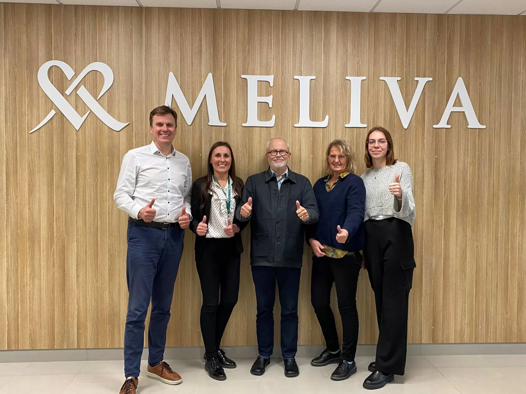 Meliva“ darbuotojų komanda stovi prie sienos su logotipu ir šypsodamiesi rodo pakeltus nykščius