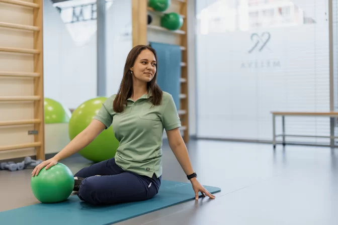 Kineziterapeutė sėdi ant kilimėlio su gimnastikos kamuoliu po treniruotės
